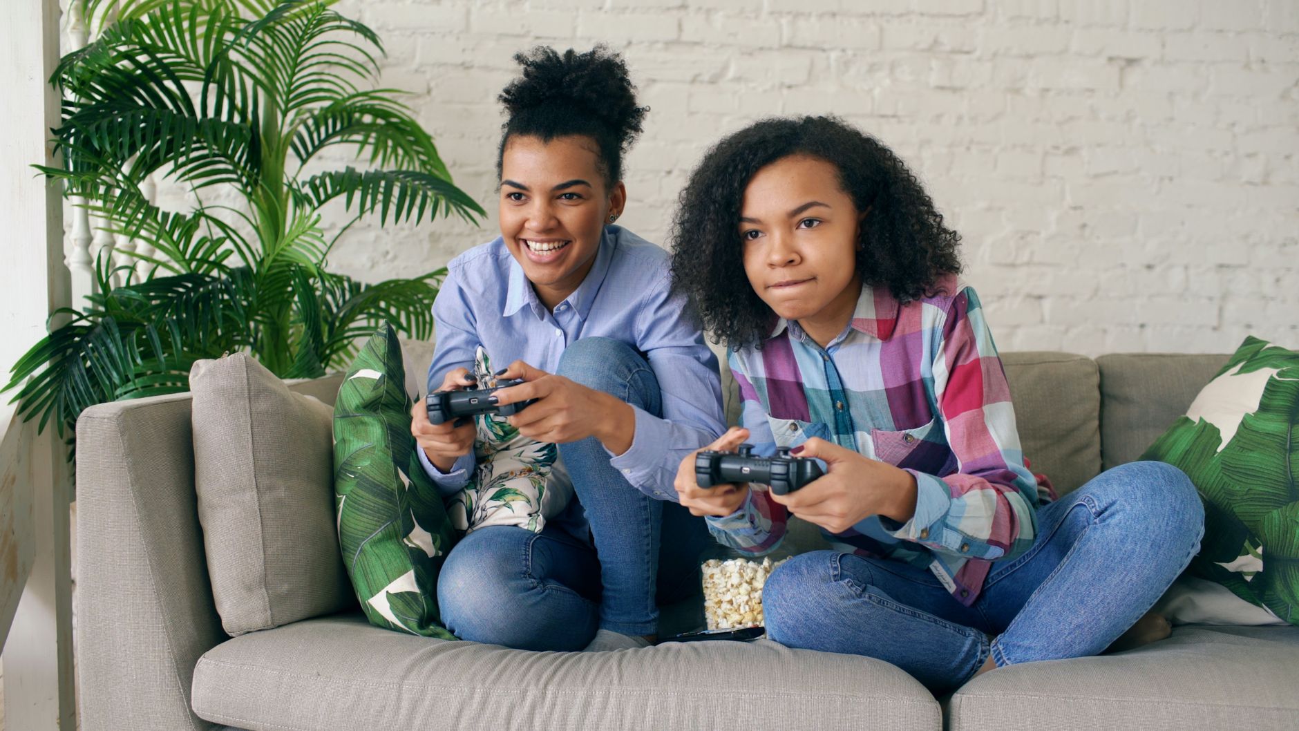 Two teenagers playing video games together on a comfortable couch, surrounded by indoor plants. — Use a Yesbet Registration C