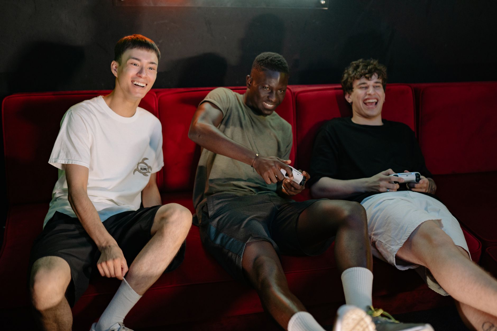 Three diverse young men sitting on a red couch, enjoying a fun video game session together. — Why Macau Togel Attracts Enthus
