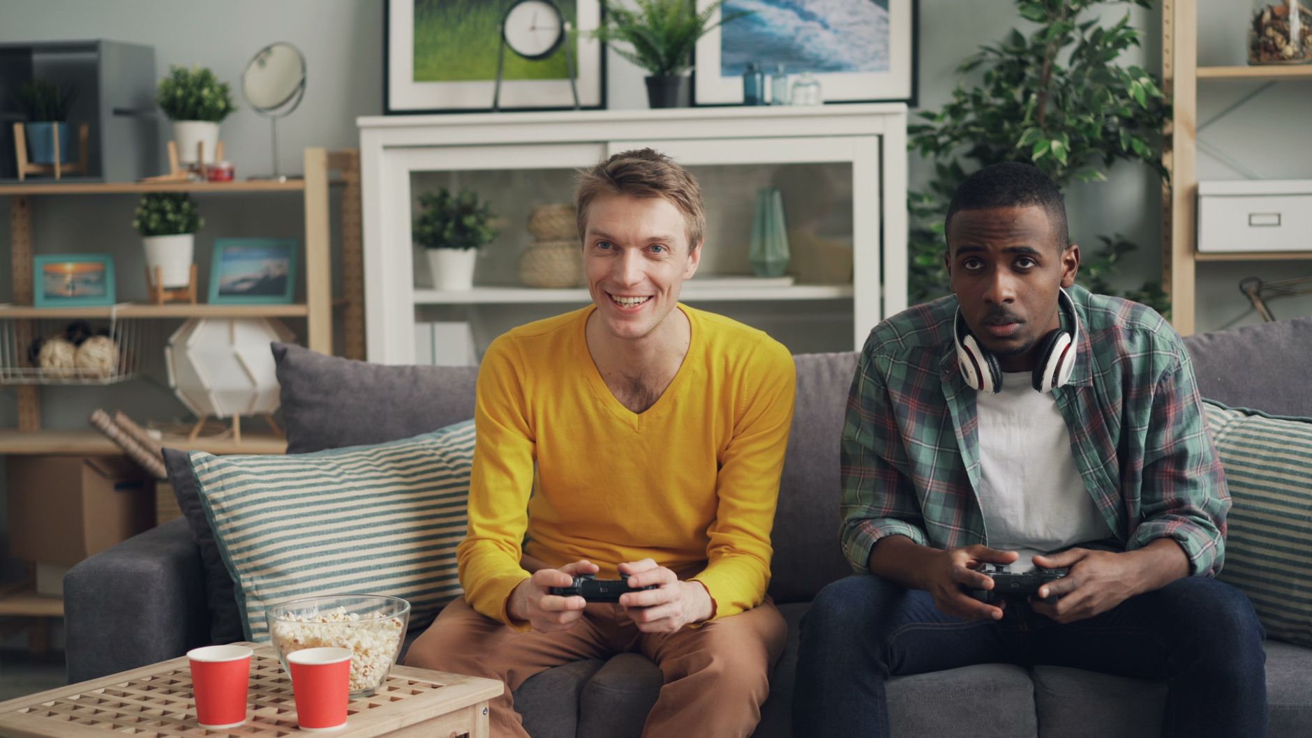 Two friends playing video games together on a sofa, one holding a controller and the other wearing headphones. — Yesbet: The